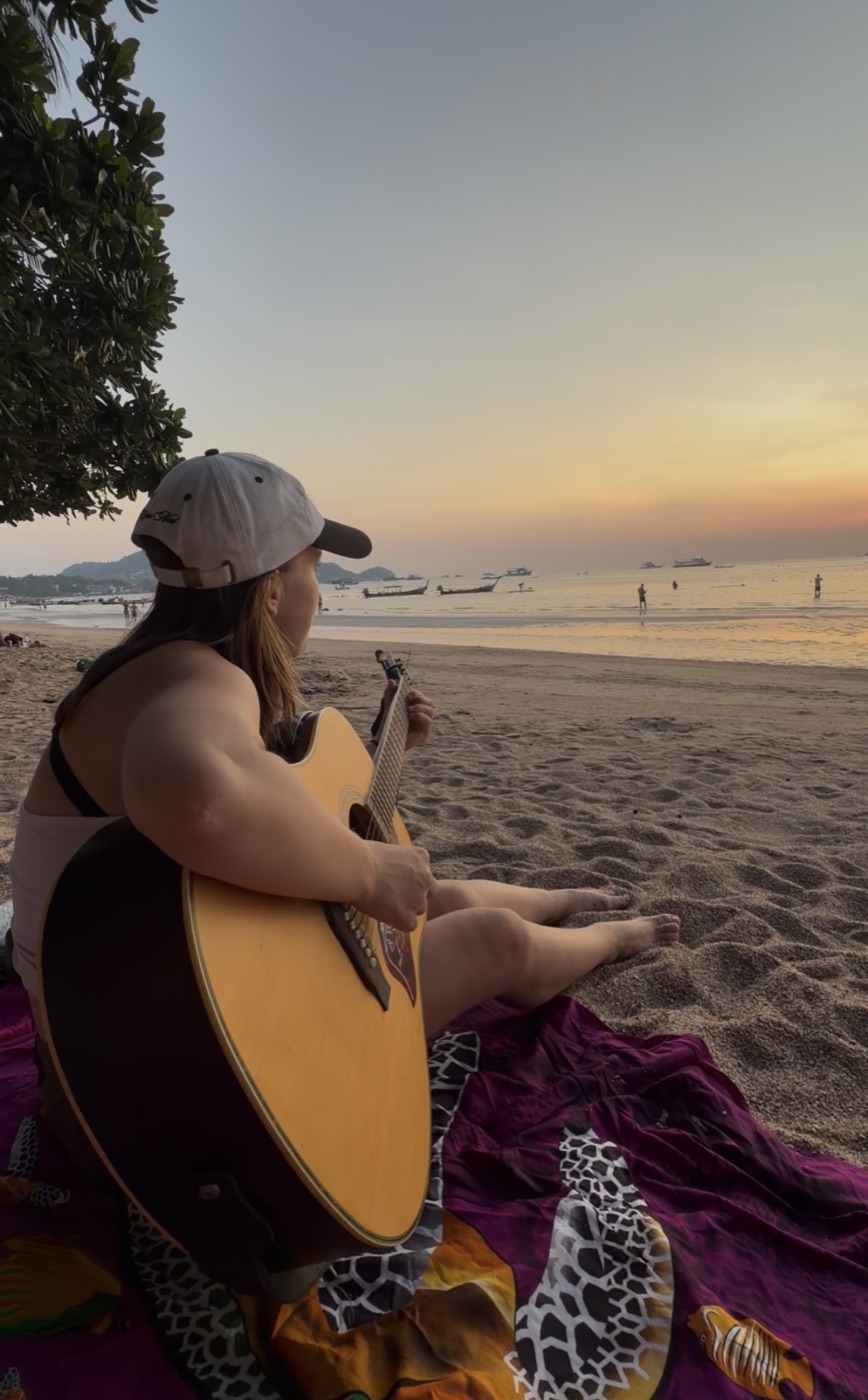 Ella Gray sitting on a beach at sunset with her guitar, looking out at the ocean